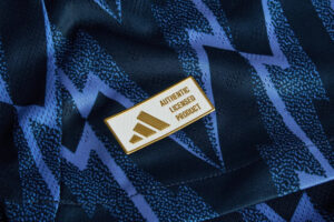 Gold rectangular pin reading 'AUTHENTIC LICENCED PRODUCT' on dark blue geometric fabric pattern.