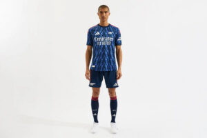 Male model wearing a blue zigzag-patterned football jersey with matching navy shorts and long navy socks, standing against a white background.