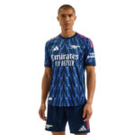 Male model wearing a blue Arsenal football away jersey with a zigzag pattern and Emirates Fly Better sponsor.