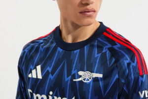 Man wearing a navy blue zigzag-pattern soccer jersey with red shoulder stripes and white logos.