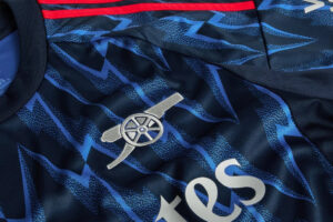 Close-up of a navy blue-patterned football jersey featuring the silver Arsenal cannon crest and partial sponsor text.