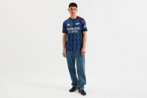 Male model wearing a blue zigzag-pattern Arsenal football jersey with 'Emirates Fly Better', jeans, and dark sneakers.