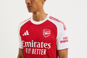 Male model wearing Arsenal home football jersey: red body, white sleeves, Emirates Fly Better sponsor, team crest visible on chest.