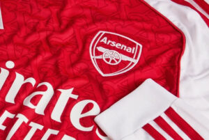 mji9536_d2 | UAE SPORT STORE Close-up of an Arsenal football jersey showing the club crest and white sponsor lettering on red fabric.