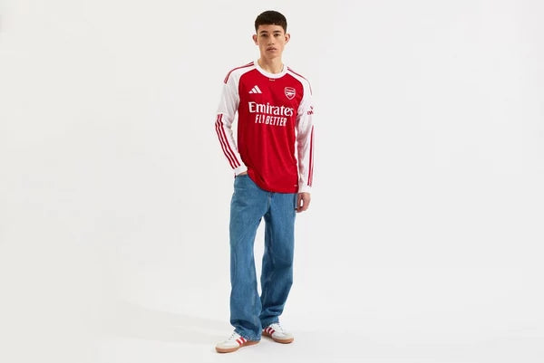 mji9536_f2 | UAE SPORT STORE Young man modeling an Arsenal red jersey with white sleeves, blue jeans, and casual sneakers, standing against a white studio background.