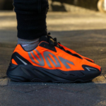 Close-up of a neon orange and black running shoe on a gray socked foot standing on pavement.
