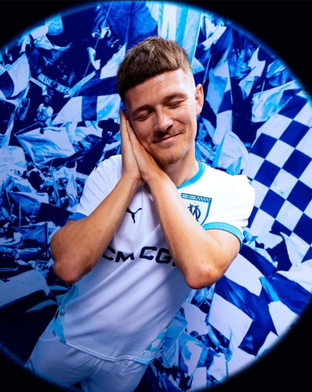 Man in a light blue Marseille jersey rests his head on hands with a content smile amid blue confetti background (circular vignette).