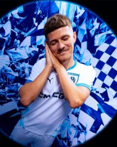 Man in a light blue Marseille jersey rests his head on hands with a content smile amid blue confetti background (circular vignette).