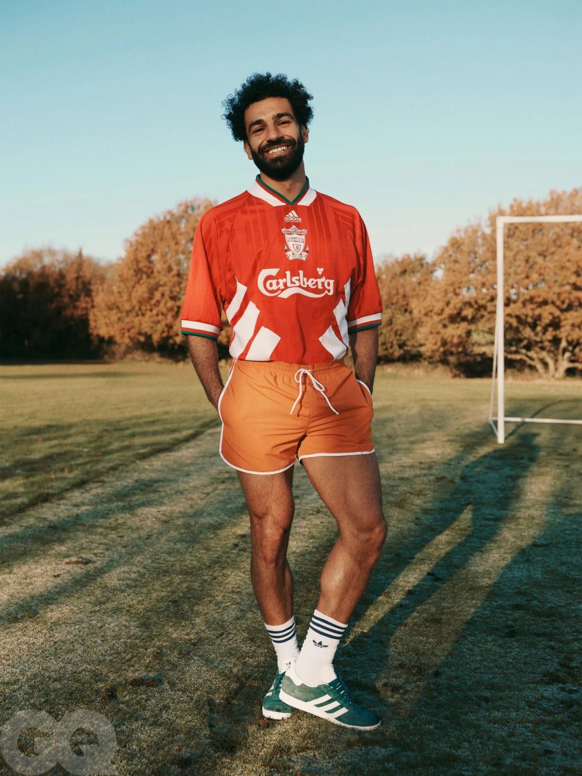 Man in a red Liverpool FC jersey poses on a grassy field, smiling with a goalpost in the background.