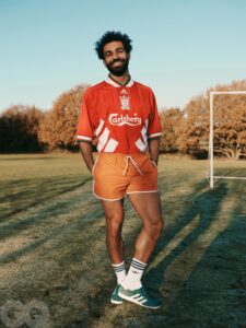 Man in a red Liverpool FC jersey poses on a grassy field, smiling with a goalpost in the background.