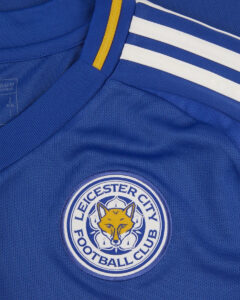 Close-up of a blue Leicester City football jersey showing the club crest with a yellow fox head and white stripes on the shoulder area.