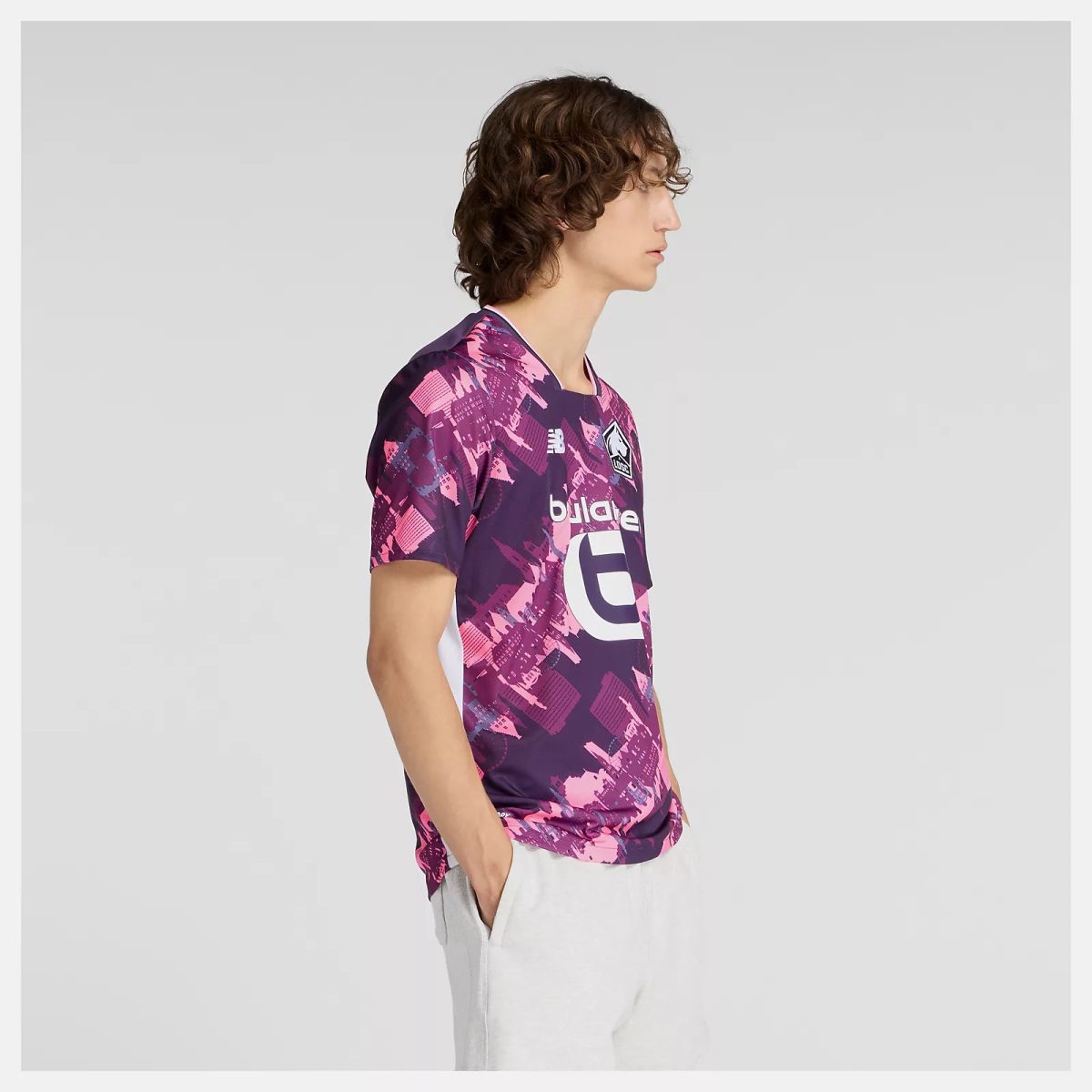 Side view of a young man in a purple-pink graphic sports jersey and light gray shorts against a neutral background.