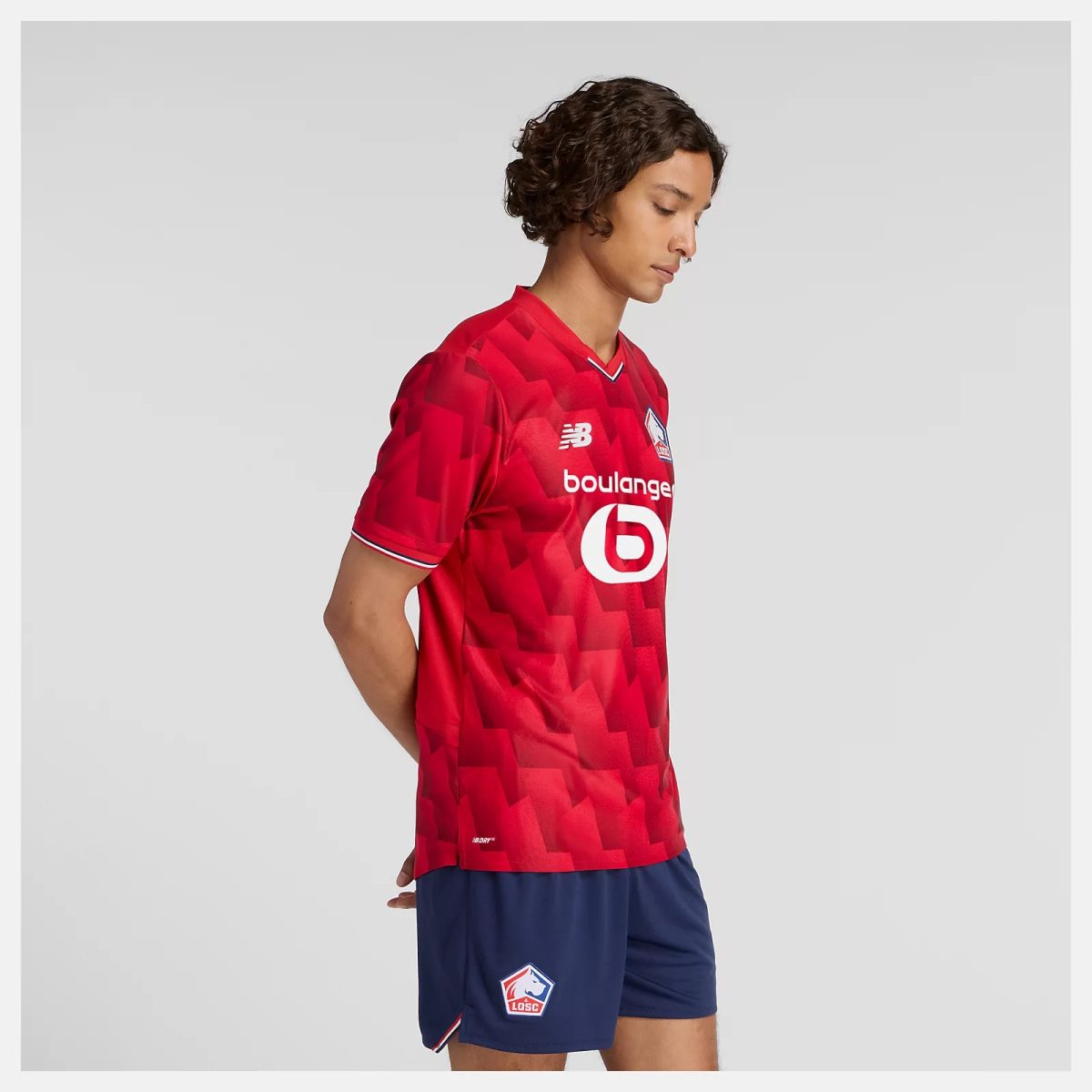 Side profile of a young man wearing a red geometric-patterned Lille OSC soccer jersey with New Balance logo and crest against a light gray background.