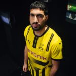 Portrait of a man with a beard wearing a yellow Borussia Dortmund jersey, looking at the camera in a dark setting, with team crest visible on chest.