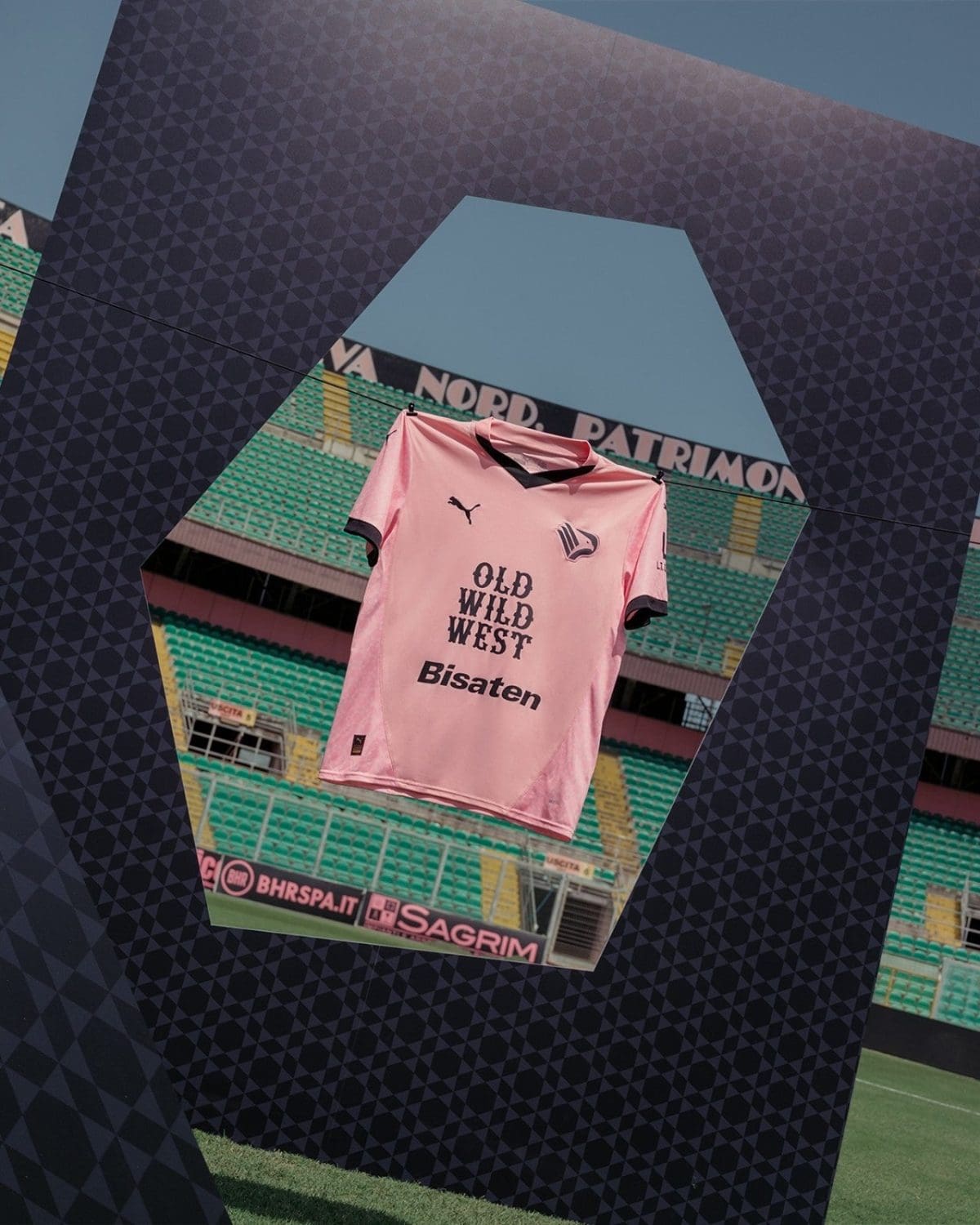 Pink football jersey with 'OLD WILD WEST' and sponsor logos hangs in a stadium, framed by a dark geometric structure.