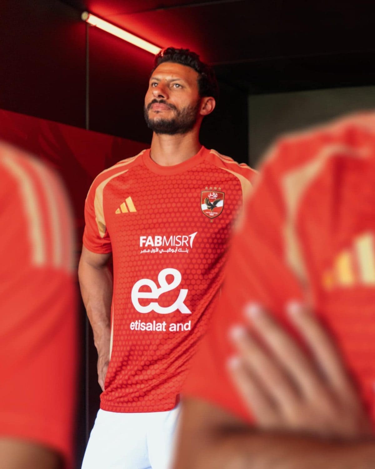 Male soccer player in a red jersey standing in a dim hallway, looking upward; team crest and sponsor logos visible on the chest.