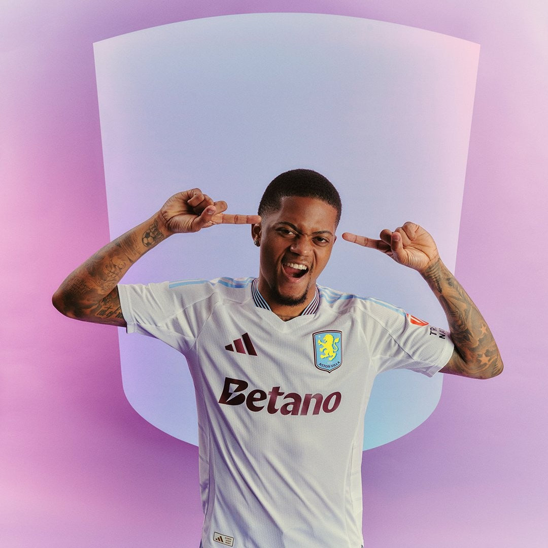 nJNRepXQNZtOwT3 | UAE SPORT STORE Soccer player in a white Aston Villa jersey raises both hands to his ears, posing against a pink-purple studio backdrop with a curved light panel behind him.