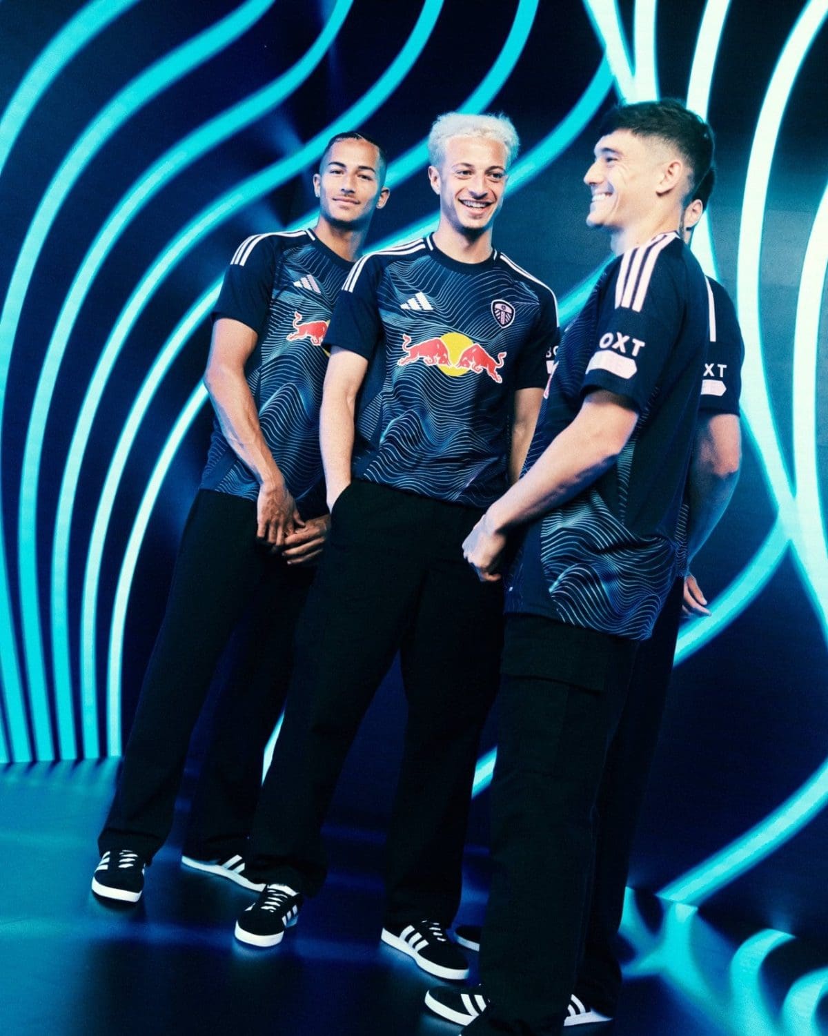 Three teammates in matching blue jerseys with Red Bull logos pose together, smiling against a neon blue, wave-pattern backdrop.