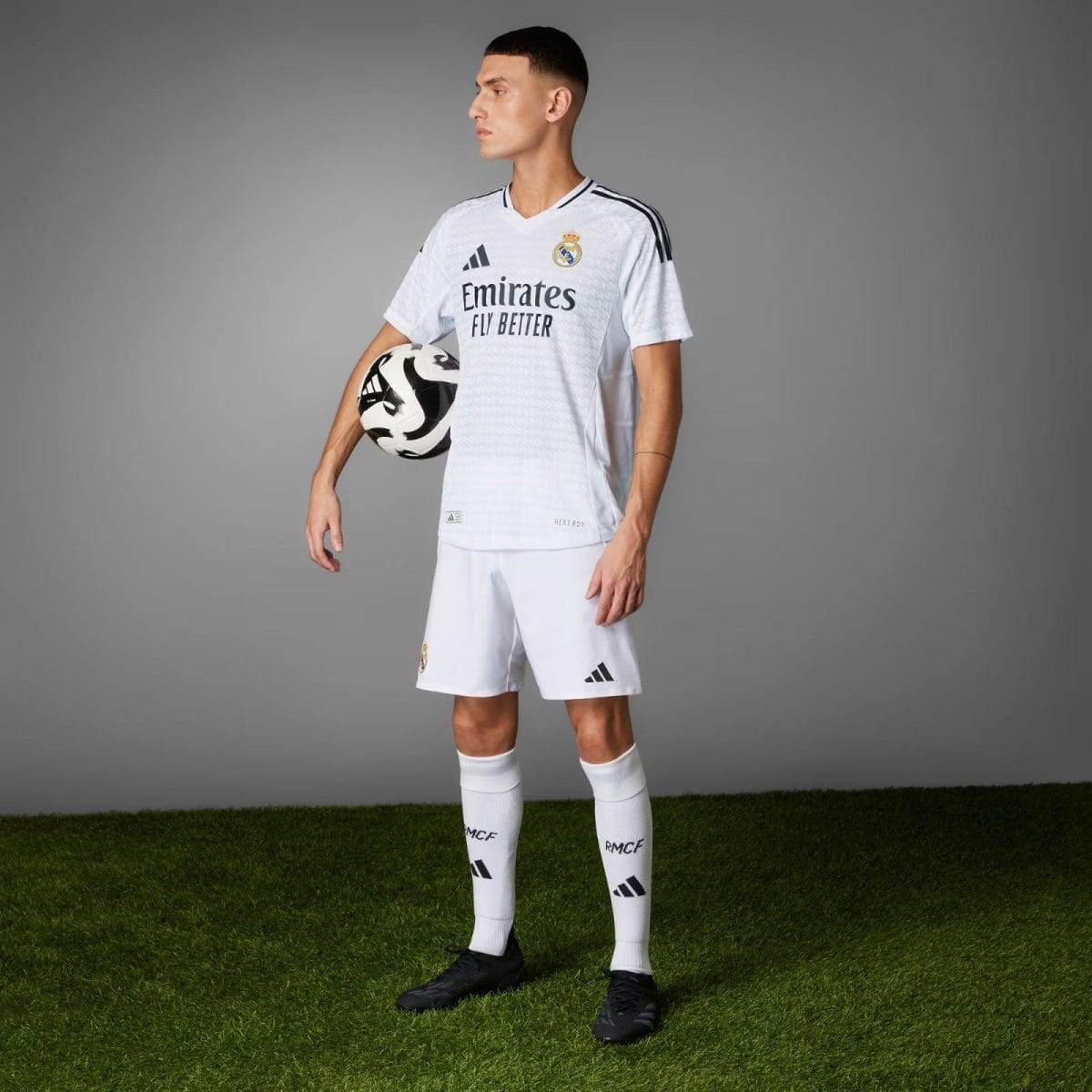 Soccer player in a white Real Madrid kit holding a black-and-white ball under his arm, standing on grass against a gray backdrop.