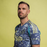 Man poses in an Ajax football jersey with yellow Ziggo sponsor against a pale yellow background.