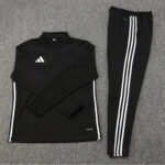 Black Adidas tracksuit top and matching pants with white side stripes laid on a gray carpeted surface.