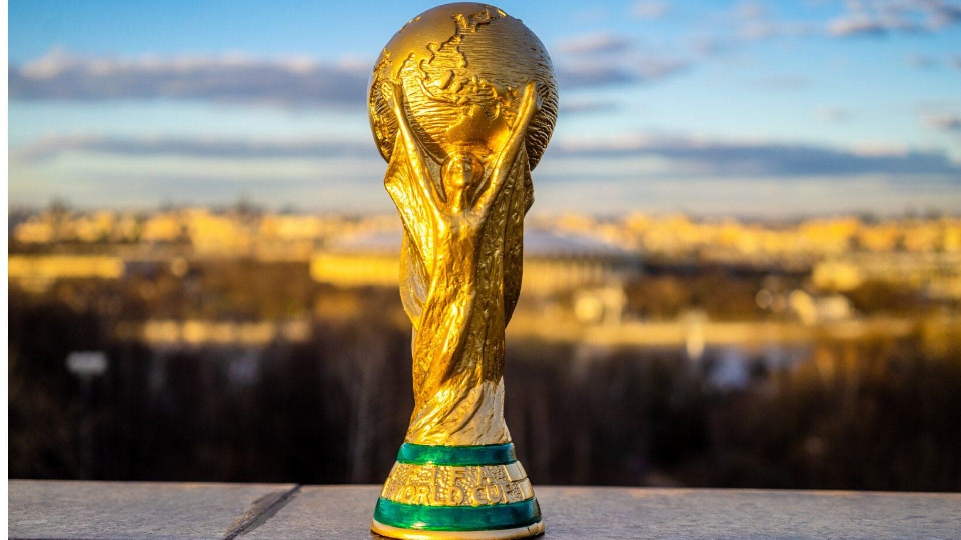 Gold FIFA World Cup trophy on a pedestal with a blurred cityscape in the background.