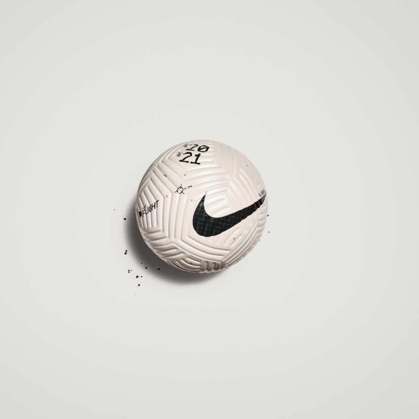 White soccer ball with a black Nike swoosh on a light gray background, small black specks nearby on the ground