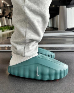 Close-up of teal, slide-style athletic shoes with a dotted sole and a small white Nike swoosh, worn with gray socks on a treadmill.