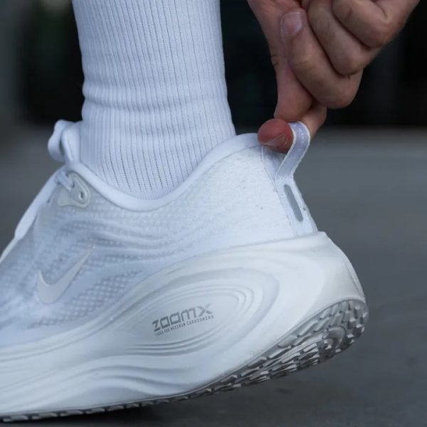 Person slides on a white Nike running shoe, pulling the heel loop to adjust the fit.