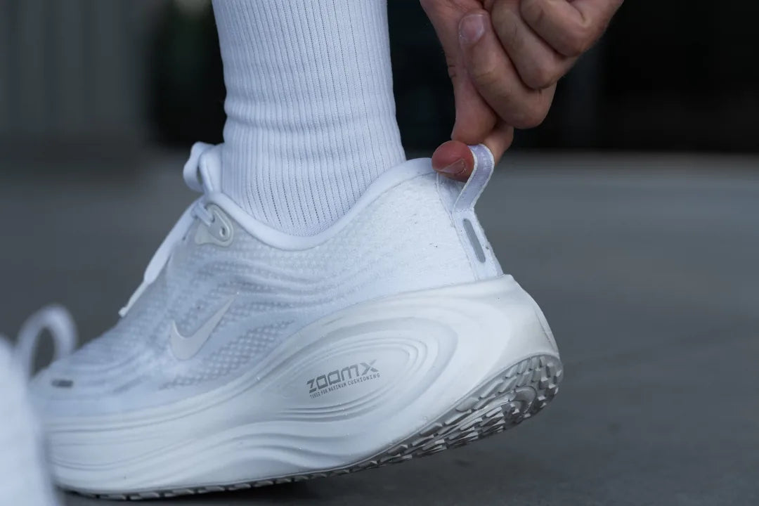 nike-vomero-plus-heel-tab-23479845-1080 | UAE SPORT STORE Person slides on a white Nike running shoe, pulling the heel loop to adjust the fit.