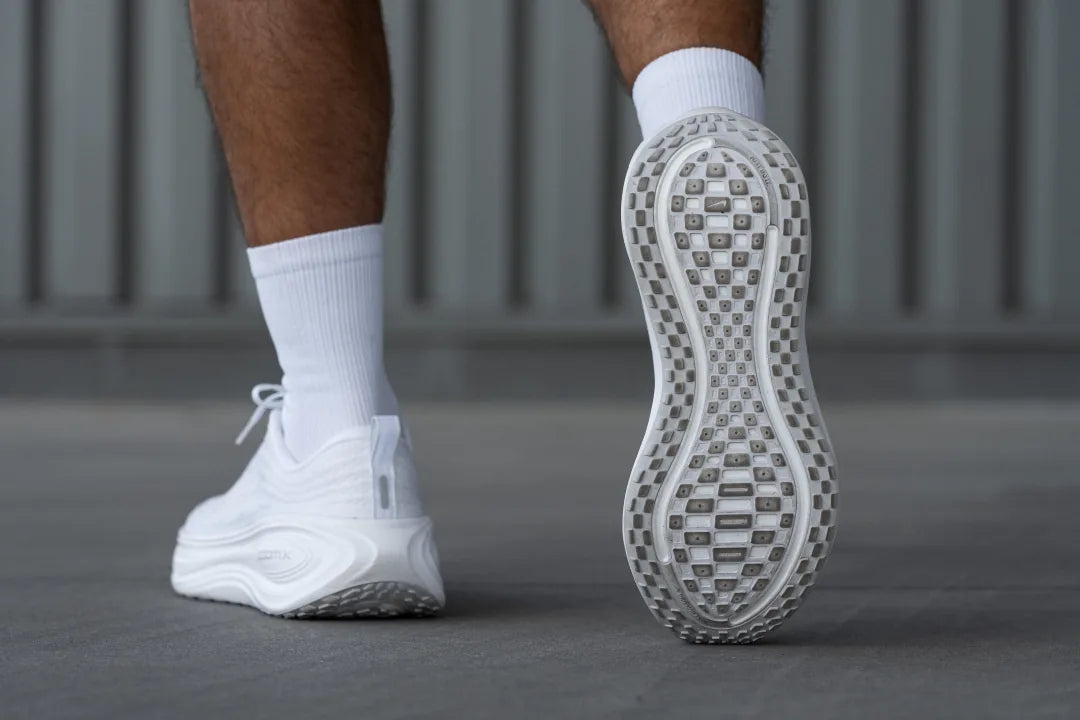 nike-vomero-plus-outsole-design-23479844-1080 | UAE SPORT STORE Person wearing white running shoes and socks, walking on gray pavement; sole tread visible.