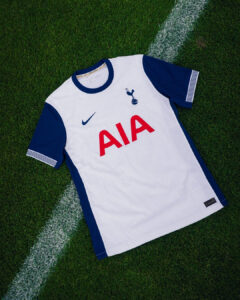 White Tottenham Hotspur jersey with blue sleeves on a grassy field displaying the AIA sponsor logo and club crest