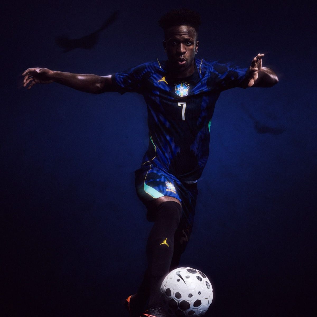 Brazilian soccer player in a blue kit with number 7 chasing a white soccer ball against a dark background, arms outstretched in motion