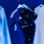Blue Manchester United jersey on a hanger in a locker area, showing Adidas stripes and Snapdragon sponsor logo.