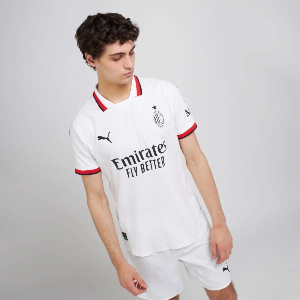 Male model wearing a white AC Milan football jersey with Emirates Fly Better sponsor, looking down to his left.