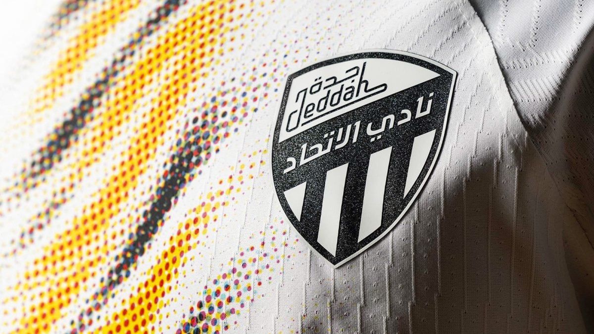 Close-up of a white soccer jersey featuring the Al-Ittihad crest (Arabic text) and black-striped shield badge.