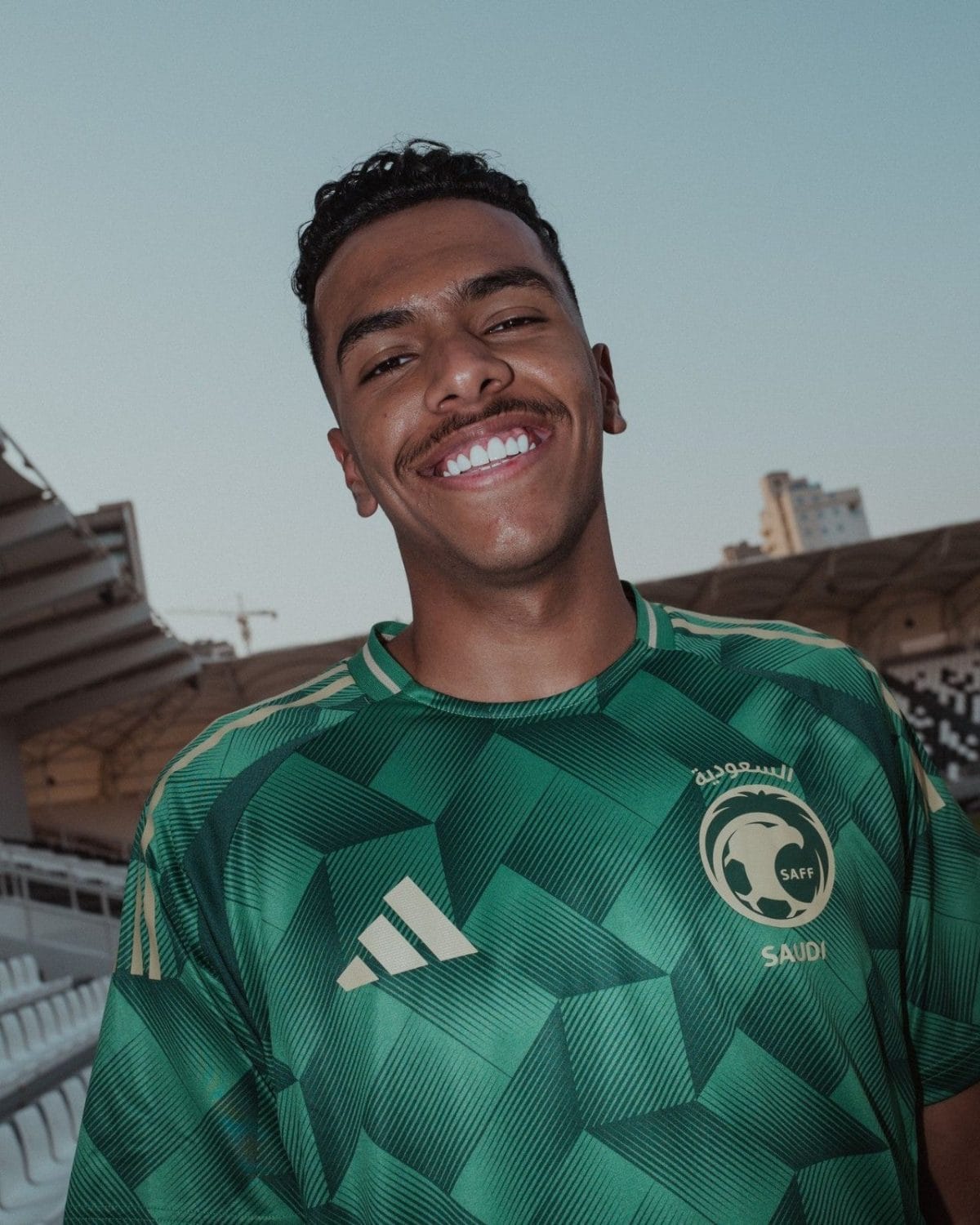 Smiling man wearing a green Saudi football jersey outdoors at a stadium backdrop.