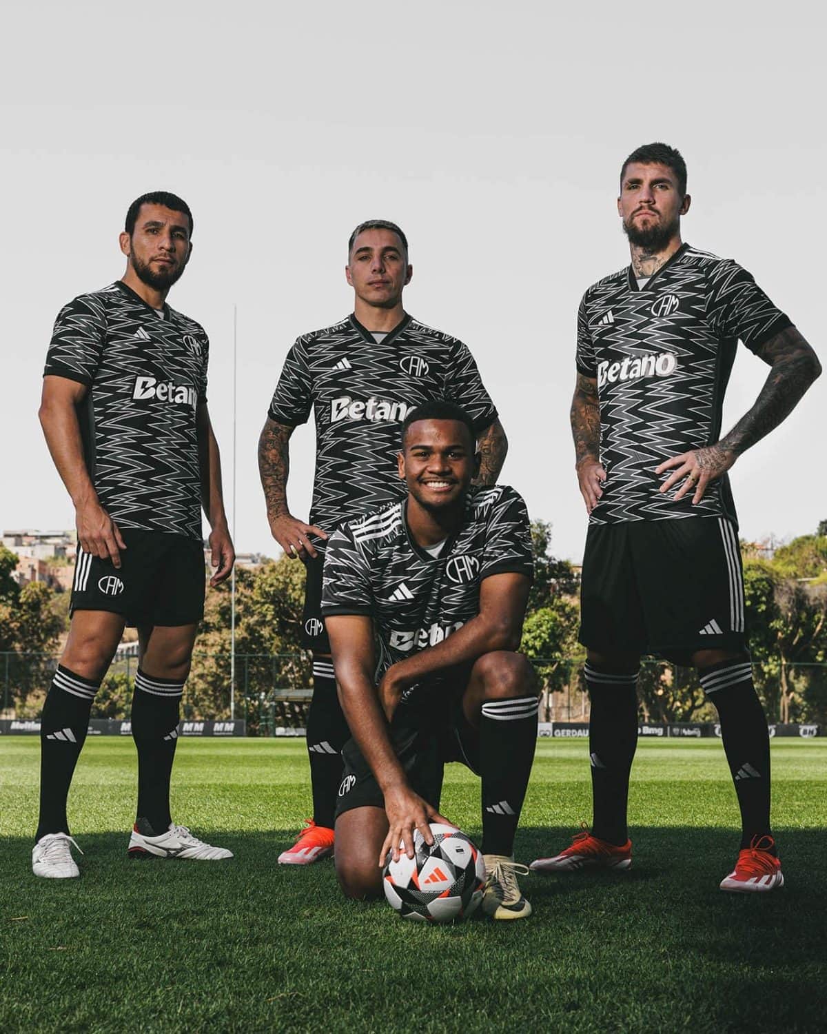 Four male soccer players pose on a training field in matching black zigzag-pattern jerseys with Betano logo; one kneels at the front with a soccer ball while three stand behind him.
