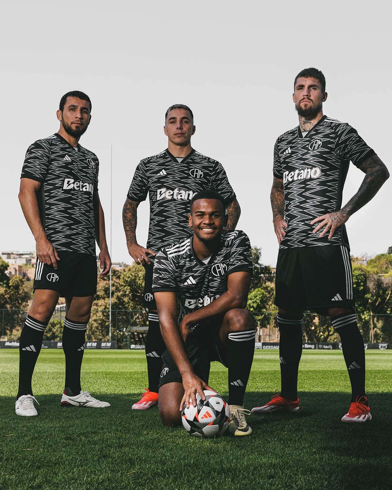 oFN3w4igVUU2IcM | UAE SPORT STORE Four male soccer players pose on a training field in matching black zigzag-pattern jerseys with Betano logo; one kneels at the front with a soccer ball while three stand behind him.