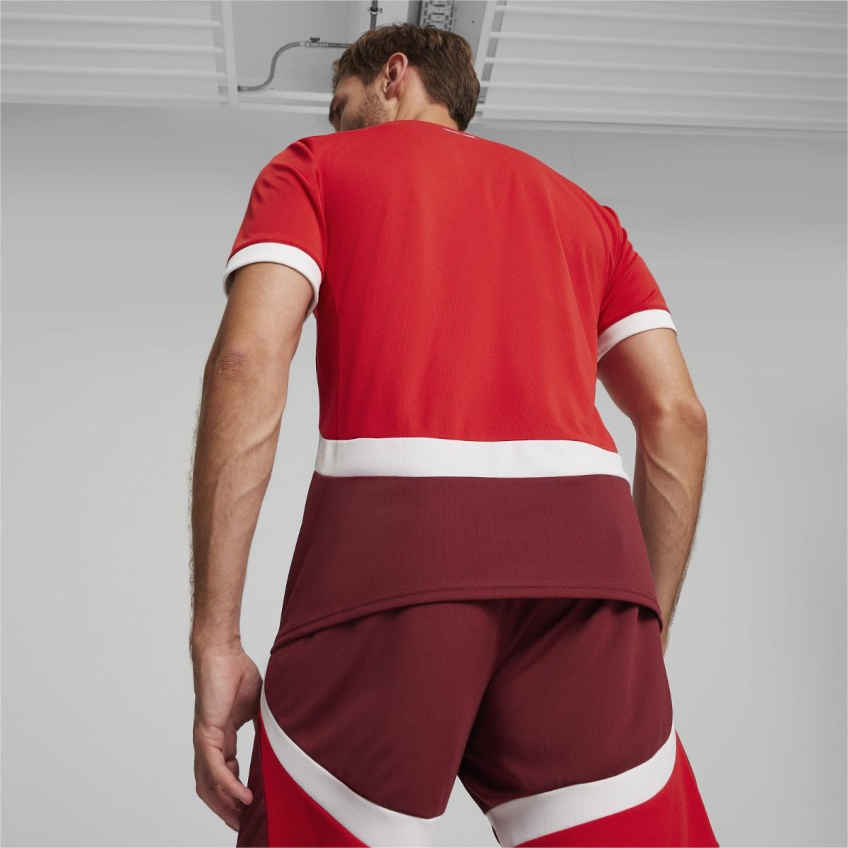 Back view of a man in a red athletic jersey with white cuffs and white diagonal stripes, standing in a neutral studio.