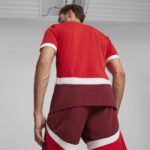 Back view of a man in a red athletic jersey with white cuffs and white diagonal stripes, standing in a neutral studio.