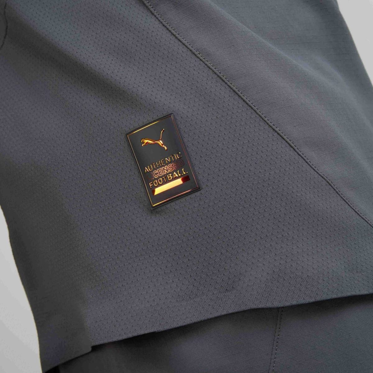 Close-up of dark textured fabric with a small gold badge showing a leaping Puma and the words 'Authentic Licensed Football'