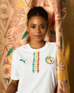 Portrait of a young woman with natural hair in a white shirt featuring colorful vertical embroidery and a circular crest, smiling softly in front of patterned fabric.
