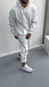 Full-body shot of a man in a white Nike hoodie and matching joggers with black piping, standing in a light room wearing white sneakers.