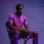 Athlete in a purple soccer kit sits on a folding chair, gazing at the camera against a purple studio backdrop.