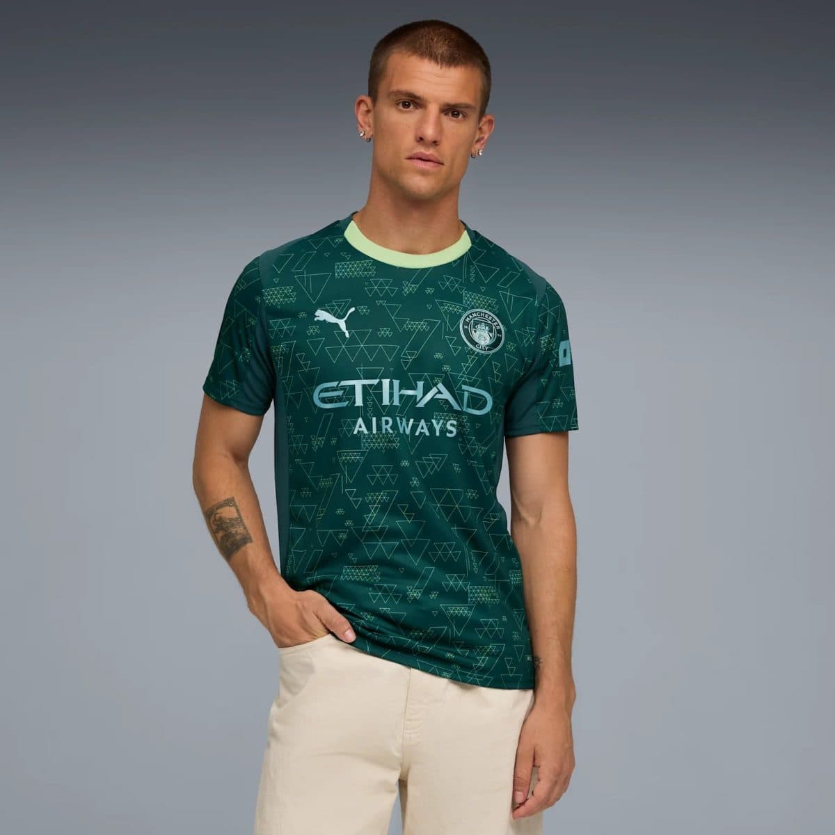 Male model wearing a teal Manchester City jersey with Etihad Airways branding, standing against a gray gradient background.