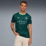 Male model wearing a teal Manchester City jersey with Etihad Airways branding, standing against a gray gradient background.