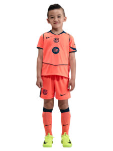Young boy in a bright orange FC Barcelona soccer kit, standing and smiling with neon yellow cleats on a white background.