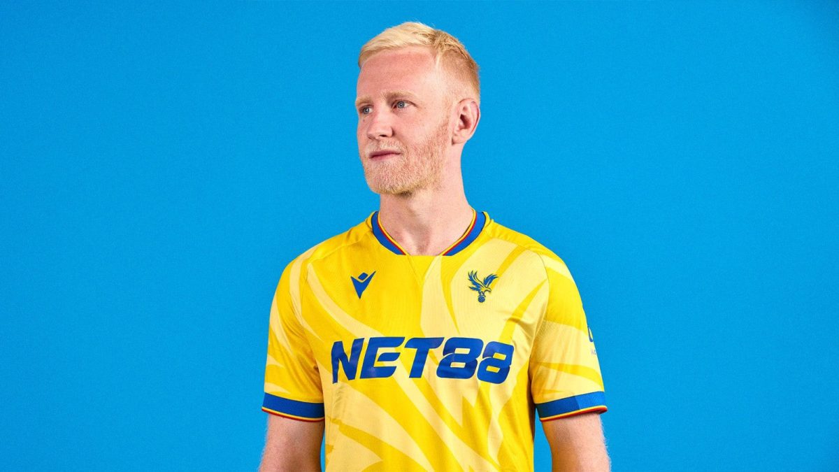 Professional soccer player standing against a bright blue background, wearing a yellow kit with NET88 sponsor.