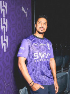 Man in a purple sports jersey standing against a purple wall with logos, looking at the camera with a calm expression.